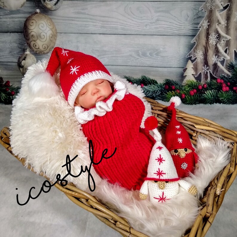 Pode incluir: Um chap&eacute;u de Papai Noel de croch&ecirc; vermelho e branco e um conjunto combinando para um beb&ecirc;, com dois gnomos de croch&ecirc; branco com detalhes vermelhos. O beb&ecirc; est&aacute; dormindo em uma cesta de vime forrada com pele branca.
