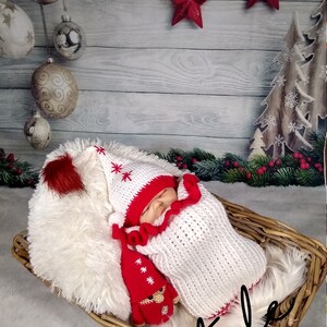Pode incluir: Um conjunto de beb&eacute; de malha branca e vermelha com um chap&eacute;u a condizer e um pompom vermelho. O conjunto &eacute; decorado com flocos de neve brancos. O beb&eacute; est&aacute; a dormir num cesto de vime com um forro de pele sint&eacute;tica branca. O fundo &eacute; um painel de madeira branca com estrelas vermelhas e uma &aacute;rvore de Natal de madeira castanha.