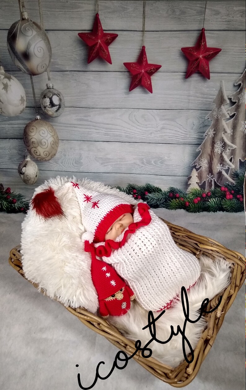 Pode incluir: Um conjunto de beb&eacute; de malha branca e vermelha com um chap&eacute;u a condizer e um pompom vermelho. O conjunto &eacute; decorado com flocos de neve brancos. O beb&eacute; est&aacute; a dormir num cesto de vime com um forro de pele sint&eacute;tica branca. O fundo &eacute; um painel de madeira branca com estrelas vermelhas e uma &aacute;rvore de Natal de madeira castanha.