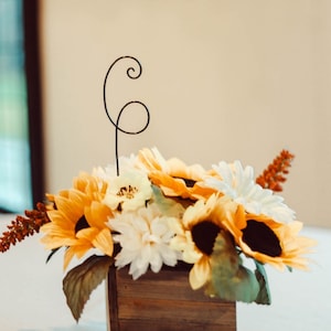 May include: A wooden box centerpiece with a floral arrangement of sunflowers, white flowers, and greenery. A black wire number "6" is placed in the arrangement.