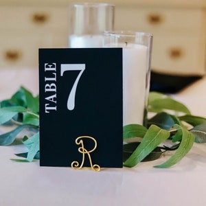 May include: Black table number card with white text that reads "TABLE 7". The card is held by a gold wire letter R.