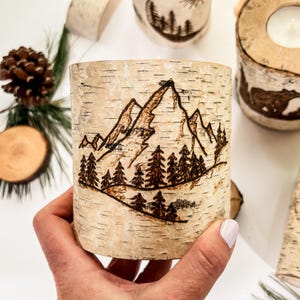 May include: A cylindrical birch bark candle holder featuring a wood-burned design of a mountain range and a forest of pine trees. The natural light-colored bark has horizontal dark markings and a rough texture, showcasing a detailed landscape scene.