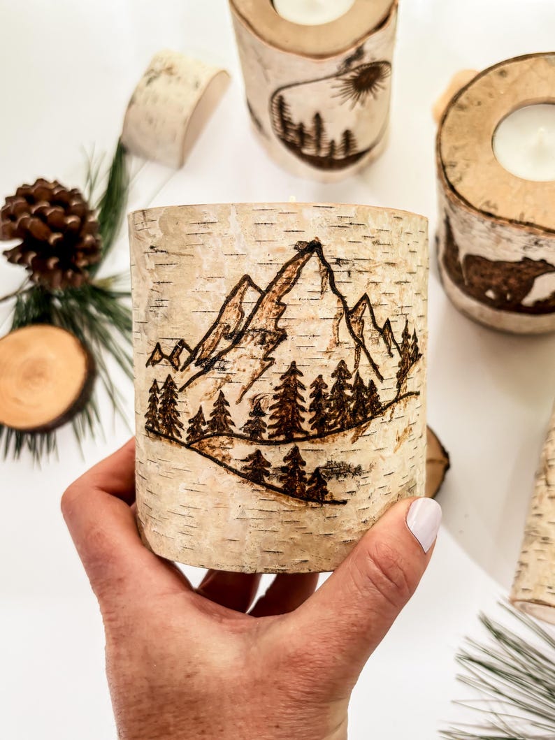 May include: A cylindrical birch bark candle holder featuring a wood-burned design of a mountain range and a forest of pine trees. The natural light-colored bark has horizontal dark markings and a rough texture, showcasing a detailed landscape scene.