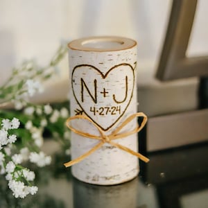 May include: A white birch wood candle holder with a heart design and the initials N+J and the date 4-27-24 engraved on it. The holder is tied with a brown ribbon.