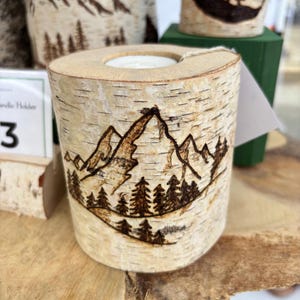 May include: A cylindrical birch wood candle holder featuring a dark brown wood-burned illustration of a mountain range and a forest of pine trees. The natural white and tan bark texture is visible on the exterior, with a circular opening at the top.