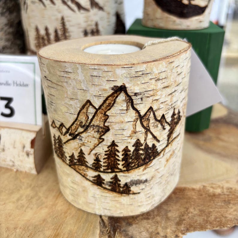 May include: A cylindrical birch wood candle holder featuring a dark brown wood-burned illustration of a mountain range and a forest of pine trees. The natural white and tan bark texture is visible on the exterior, with a circular opening at the top.