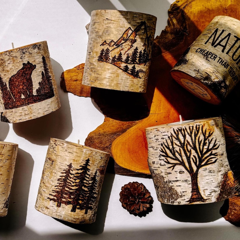 May include: Five cylindrical birch bark candles featuring wood-burned designs including a bear, a mountain landscape, a tree, and text that reads NATURE CHEAPER THAN THERAPY. Each candle has a single white wick centered on the top surface.