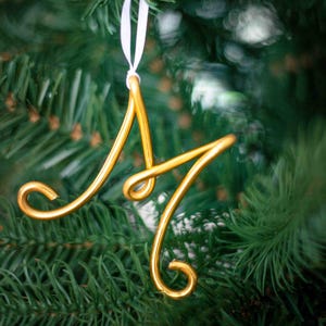 May include: A gold-colored metal ornament shaped like the letter "M" hanging on a Christmas tree.