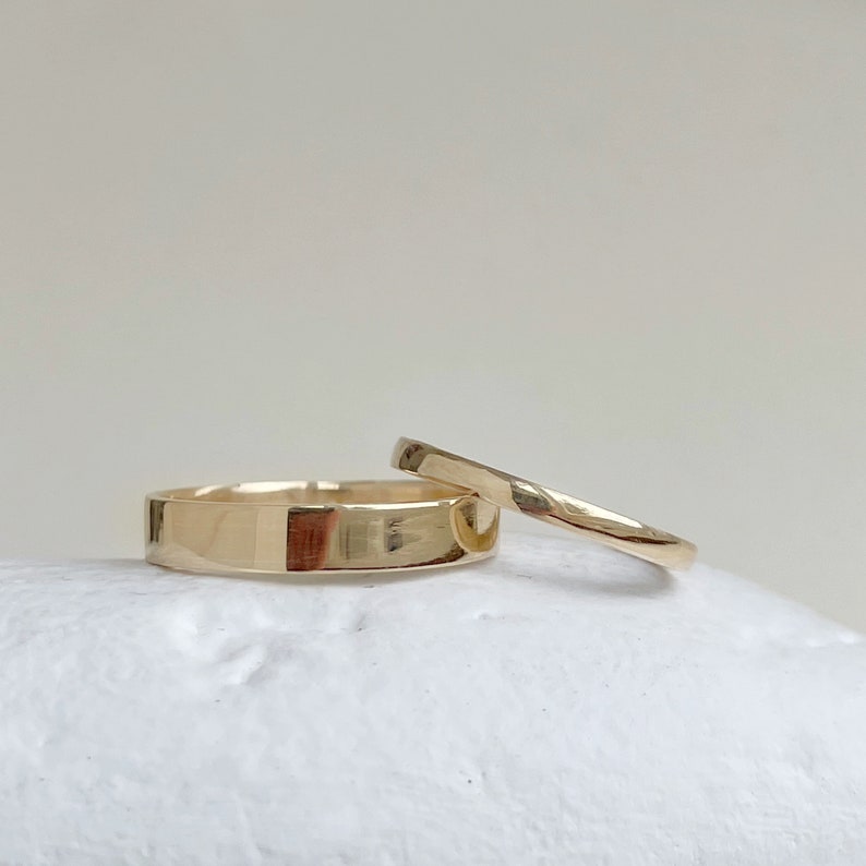 May include: Two gold wedding bands, one wider than the other, are stacked on a white surface.