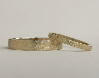 May include: Two gold wedding bands with a textured surface. One band is wider than the other. The rings are set against a neutral background, highlighting their simple elegance. The bands are likely made of gold.