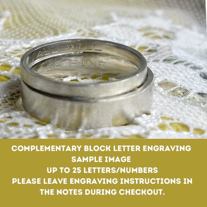 May include: Two silver wedding bands with a block letter engraving that reads "All will be fine".  The bands are on a white lace background.  The image is a sample of a custom engraving service that allows up to 25 letters or numbers.