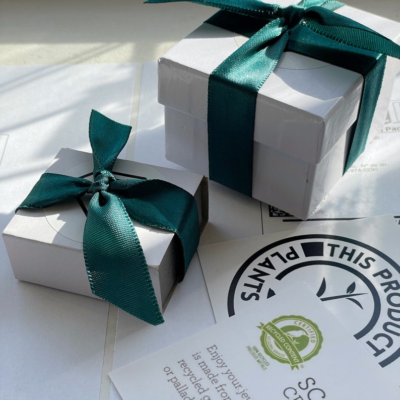 May include: Two white gift boxes with green ribbon bows. The boxes are sitting on a white surface. A sticker with the text "THIS PRODUCT IS MADE FROM PLANTS" is visible in the background.
