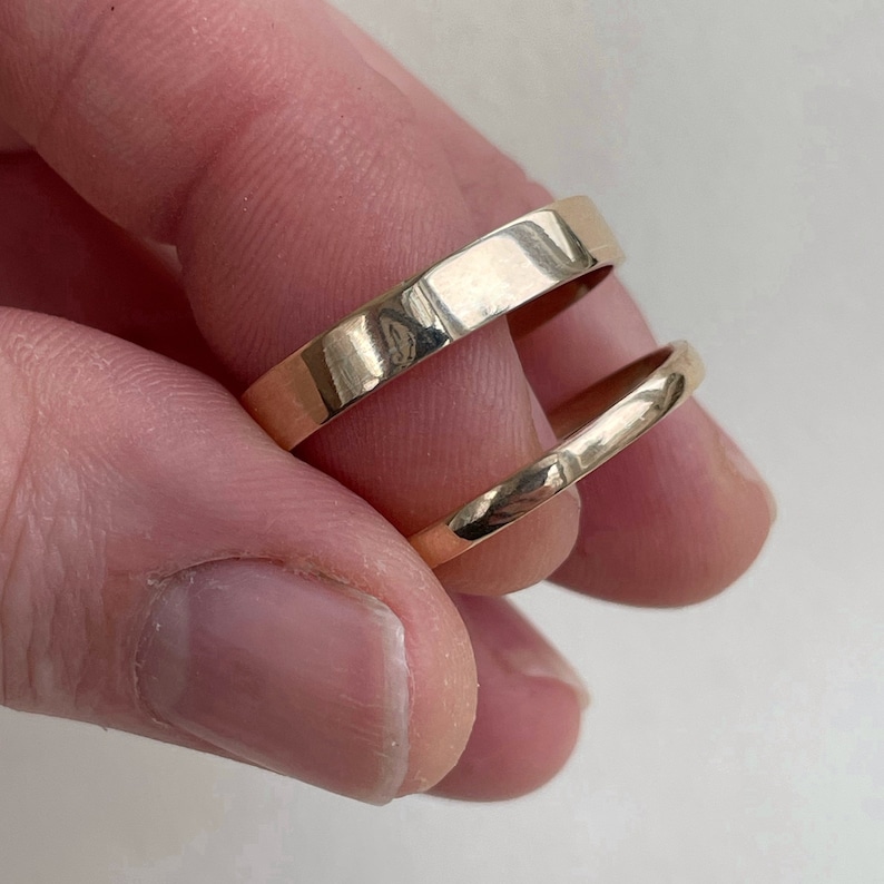 May include: Two gold-colored metal bands, one wider than the other, are stacked on top of each other. The bands are smooth and shiny.