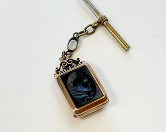 Op de afbeelding: Antieke goudkleurige horlogefob met een rechthoekige zwarte camee hanger. De hanger heeft een gedetailleerde reliëfsnede. De fob heeft een T-bar en een ketting met een kleine ovale steen. Het stuk is geplaatst tegen een witte achtergrond.