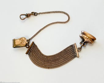 Op de afbeelding: Antieke goudkleurige horlogeketting met een decoratieve sluiting en een geweven mesh-gedeelte. De ketting bevat een draaisluiting en een decoratieve fob. Het metaal heeft een warme, verouderde uitstraling.