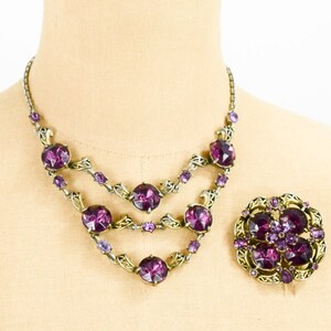 May include: A vintage purple rhinestone necklace and brooch set. The necklace has three rows of large, round rhinestones with a gold-tone metal setting. The brooch is a floral design with smaller rhinestones.