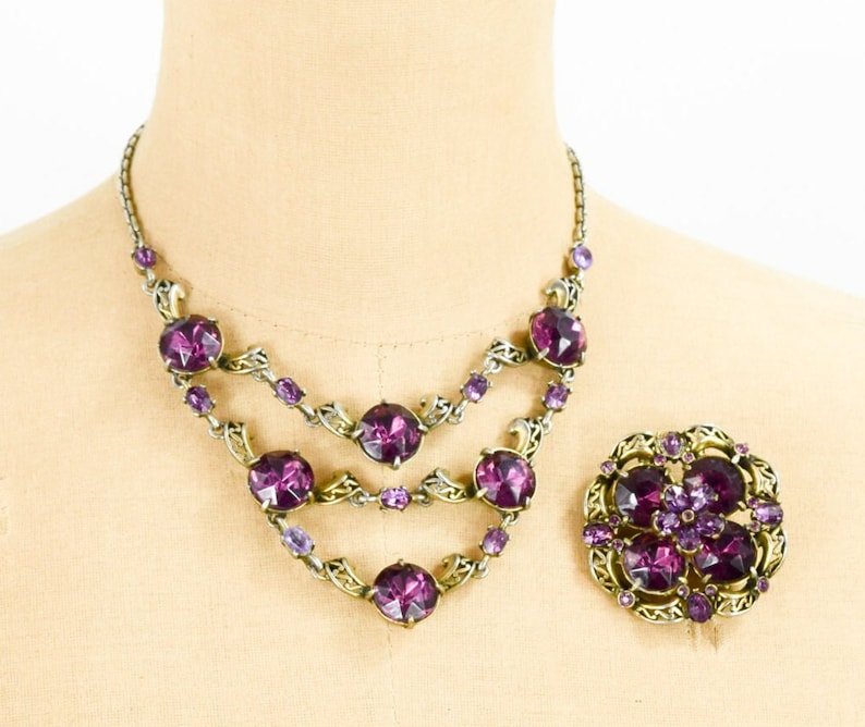 May include: A vintage purple rhinestone necklace and brooch set. The necklace has three rows of large, round rhinestones with a gold-tone metal setting. The brooch is a floral design with smaller rhinestones.