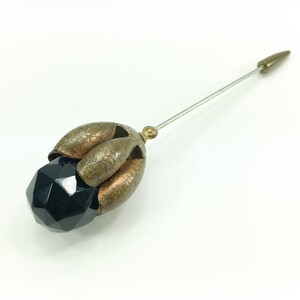 May include: A black glass bead with a faceted design is attached to a gold-toned metal pin with a pointed end.