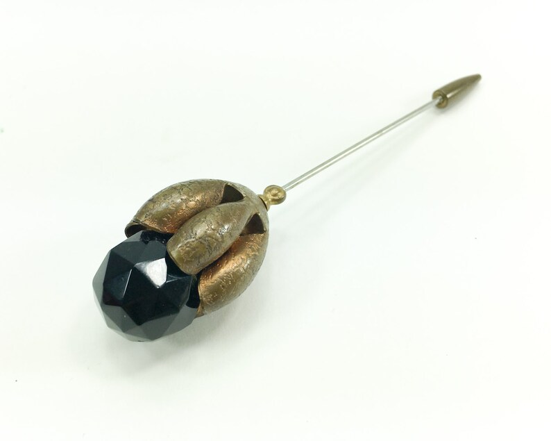 May include: A black glass bead with a faceted design is attached to a gold-toned metal pin with a pointed end.