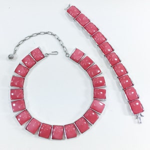 May include: A pink and silver necklace and bracelet set with square-shaped stones. The stones have a carved floral design.