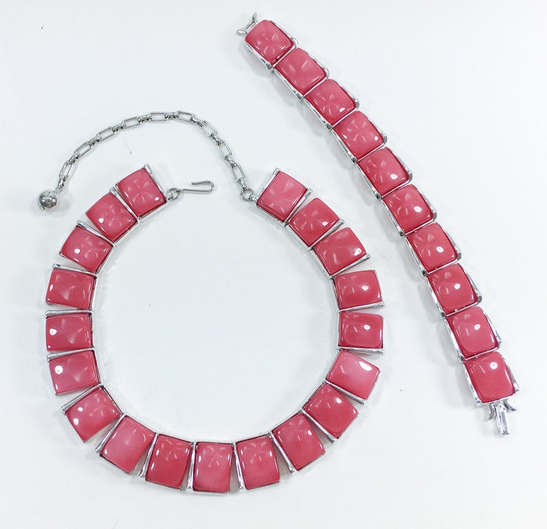 May include: A pink and silver necklace and bracelet set with square-shaped stones. The stones have a carved floral design.
