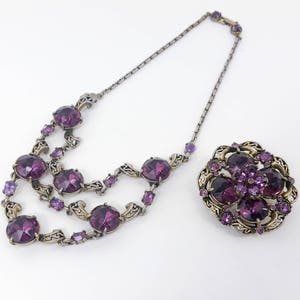 May include: A purple rhinestone necklace and brooch set with a silver tone chain and ornate metalwork.