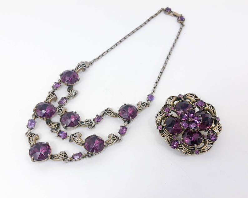 May include: A purple rhinestone necklace and brooch set with a silver tone chain and ornate metalwork.