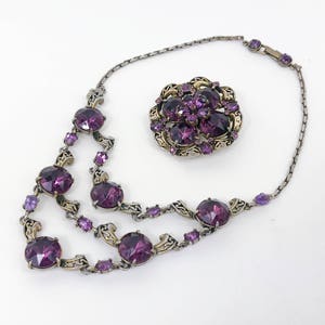 May include: A silver chain necklace with a purple rhinestone pendant and a matching brooch. The necklace has multiple strands of chain with large, round purple rhinestones.
