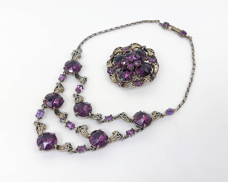 May include: A silver chain necklace with a purple rhinestone pendant and a matching brooch. The necklace has multiple strands of chain with large, round purple rhinestones.
