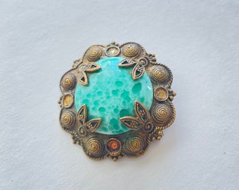 May include: A round, green, glass brooch with a textured surface. The brooch is surrounded by a gold metal frame with intricate details.