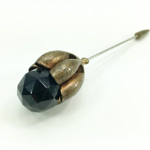 May include: A black faceted glass bead with a gold metal setting in the shape of a flower. The pin is attached to the back of the setting.