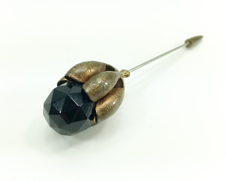 May include: A black faceted glass bead with a gold metal setting in the shape of a flower. The pin is attached to the back of the setting.