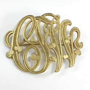 May include: A vintage gold-toned monogram brooch. The brooch features an ornate, intertwined design of three letters, possibly initials. The letters are in a flowing, Art Nouveau style, with decorative flourishes. The brooch is set against a white background.