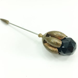 May include: A black glass bead with a faceted design is encased in a gold-toned metal setting. The setting has a flower-like shape with five petals. A long, thin metal pin extends from the back of the setting.