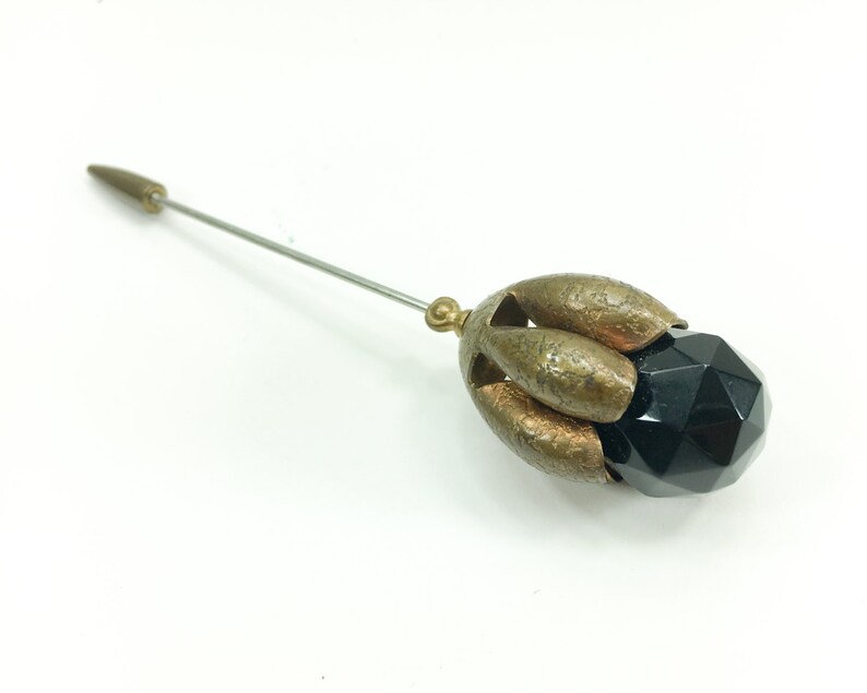 May include: A black glass bead with a faceted design is encased in a gold-toned metal setting. The setting has a flower-like shape with five petals. A long, thin metal pin extends from the back of the setting.