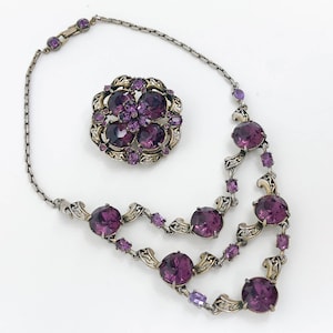 May include: A silver chain necklace with a purple rhinestone pendant and matching brooch. The necklace has a delicate design with swirls and a double row of large rhinestones.