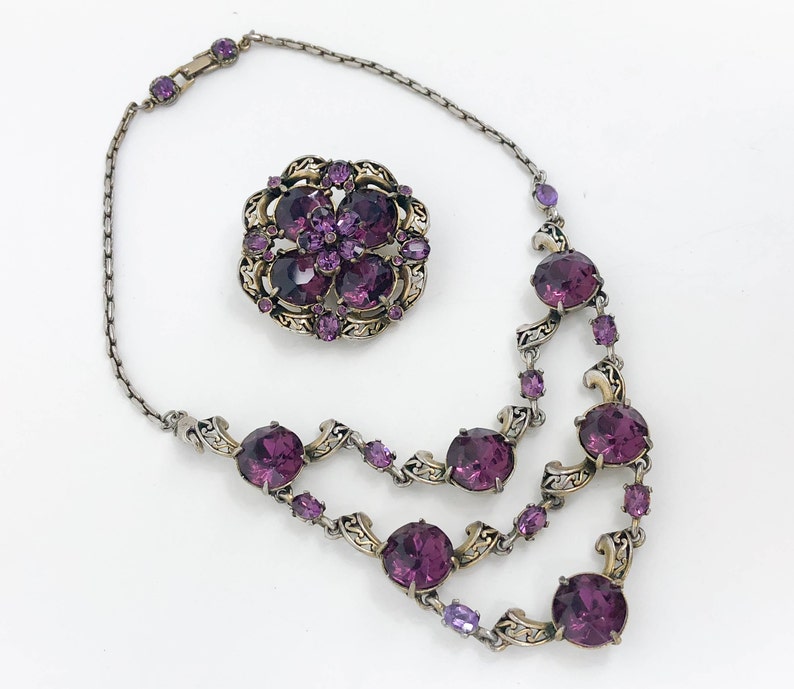 May include: A silver chain necklace with a purple rhinestone pendant and matching brooch. The necklace has a delicate design with swirls and a double row of large rhinestones.