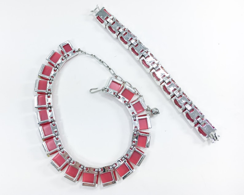 May include: A pink and silver necklace and bracelet set. The necklace and bracelet are made of linked metal squares with pink stones in the center of each square.