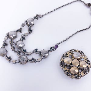 May include: A silver necklace with a silver chain and multiple rows of round, clear stones. The necklace is attached to a brooch with a gold-toned filigree design and five round, clear stones.