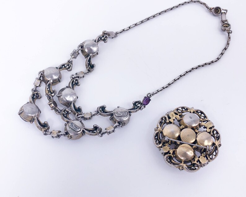 May include: A silver necklace with a silver chain and multiple rows of round, clear stones. The necklace is attached to a brooch with a gold-toned filigree design and five round, clear stones.