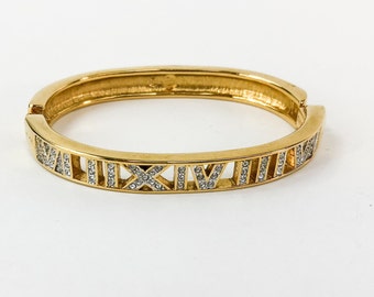 以下が含まれることがあります： ローマ数字の切り抜きとキラキラ光るクリスタルアクセントが特徴の、ゴールドトーンのヒンジ付きバングルブレスレット。普段使いにも特別な場面にも適した、クラシックなジュエリーです。