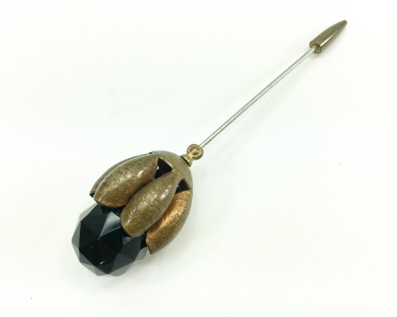 May include: A vintage black glass and brass hat pin with a pointed metal tip. The pin features a faceted black glass ball topped with a brass flower design.