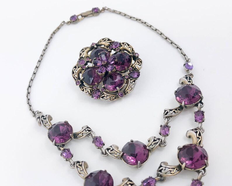 May include: A silver and gold necklace with a large purple rhinestone flower pendant and smaller purple rhinestones throughout the chain.