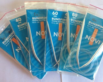 May include: Five packages of circular knitting needles in various sizes, ranging from 30 cm to 120 cm. The needles are silver and are packaged in clear plastic bags with blue and orange labels. The labels have the brand name "NOVI" and the text "RUNDPINNE" or "RUNDPIN".
