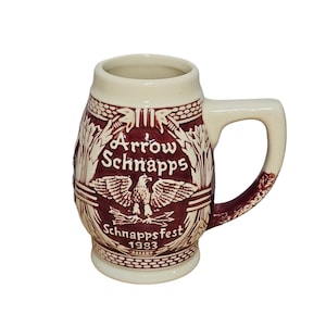May include: A white ceramic beer stein with a brown design. The stein features an eagle with outstretched wings, the text "Arrow Schnapps", and the text "Schnappsfest 1983".
