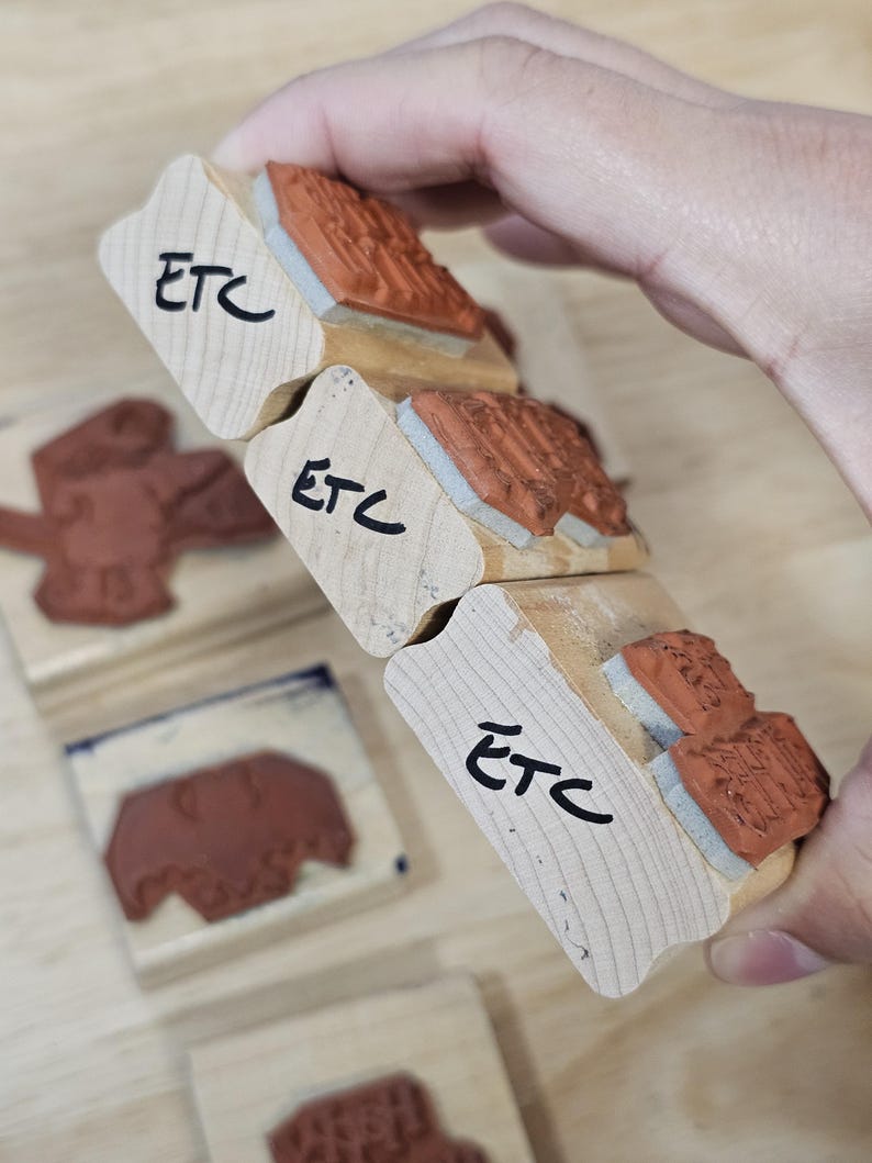 Pode incluir: Carimbos de madeira com almofadas de tinta vermelha. Os carimbos t&ecirc;m as letras "ETC" escritas em tinta preta. Os carimbos s&atilde;o segurados em uma m&atilde;o.
