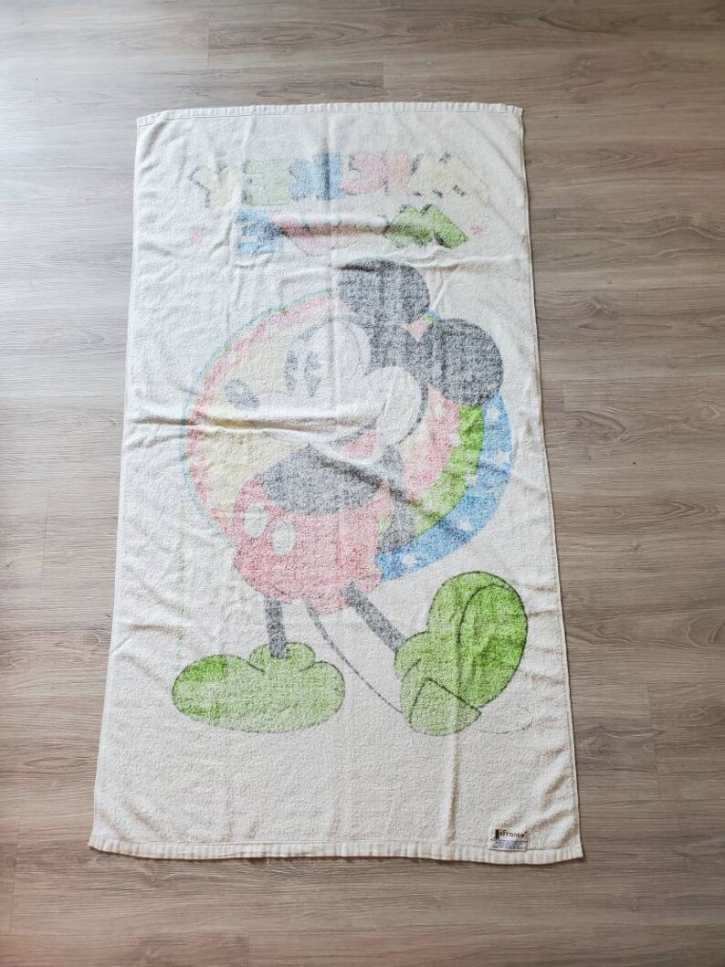 May include: A white beach towel with a colorful Mickey Mouse design. The towel features a rainbow circle around Mickey Mouse, who is wearing red shorts and green shoes. The towel has the text "Disney" printed on the bottom right corner.