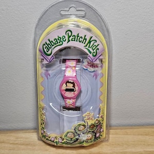 May include: A pink Cabbage Patch Kids watch with a white daisy print band. The watch face features a Cabbage Patch Kid doll with a pink dress and a white daisy on her head. The watch is packaged in a clear plastic clamshell.
