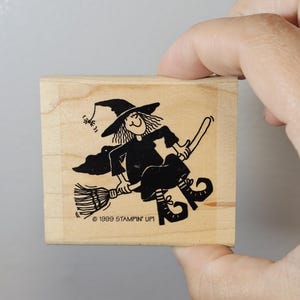 Pode incluir: Um carimbo de madeira com uma bruxa de desenho animado preta em uma vassoura. A bruxa usa um chap&eacute;u pontudo e um vestido. O texto "1999 STAMPIN' UP!" &eacute; impresso abaixo. O carimbo &eacute; segurado por uma m&atilde;o.