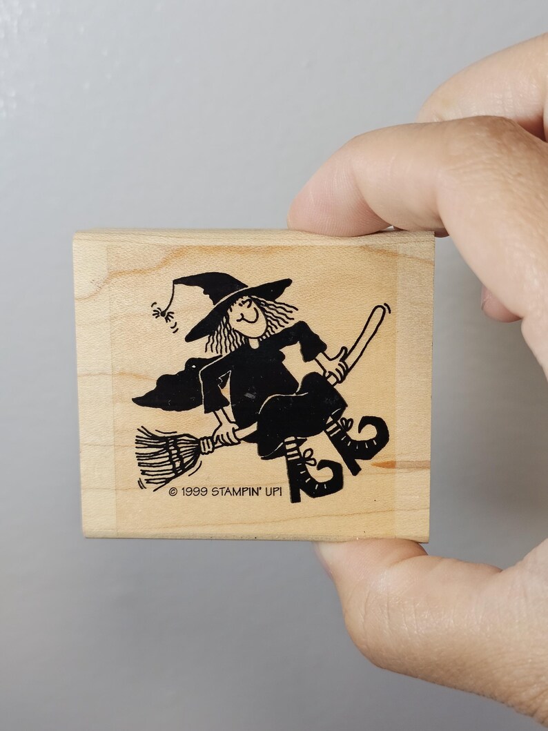 Pode incluir: Um carimbo de madeira com uma bruxa de desenho animado preta em uma vassoura. A bruxa usa um chap&eacute;u pontudo e um vestido. O texto "1999 STAMPIN' UP!" &eacute; impresso abaixo. O carimbo &eacute; segurado por uma m&atilde;o.